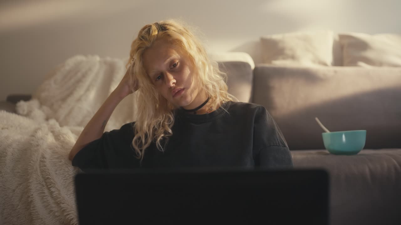 A sad blonde girl in a gray T-shirt leans on her hand and sadly looks at the laptop screen when they call her and she stands near the sofa at home