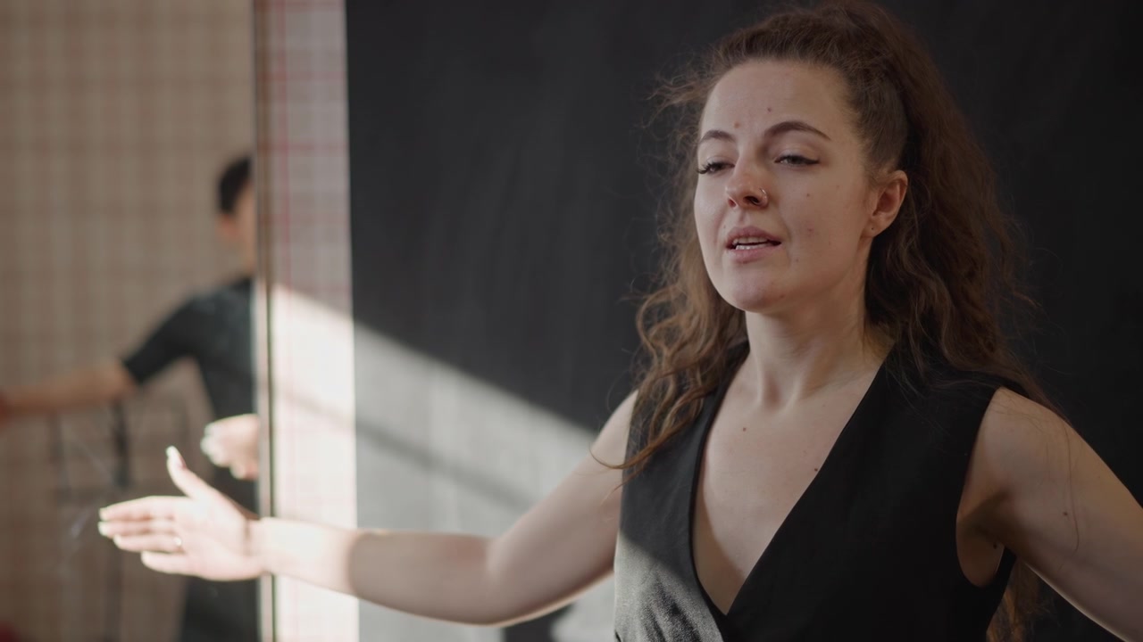 Cantante mujer de cabello rizado en sala de ensayo demostrando técnica de respiración y gestos expresivos, silueta de mentor al fondo, luz solar proyectando sombras en la pared de azulejos, retrato de cerca, aula de estudio