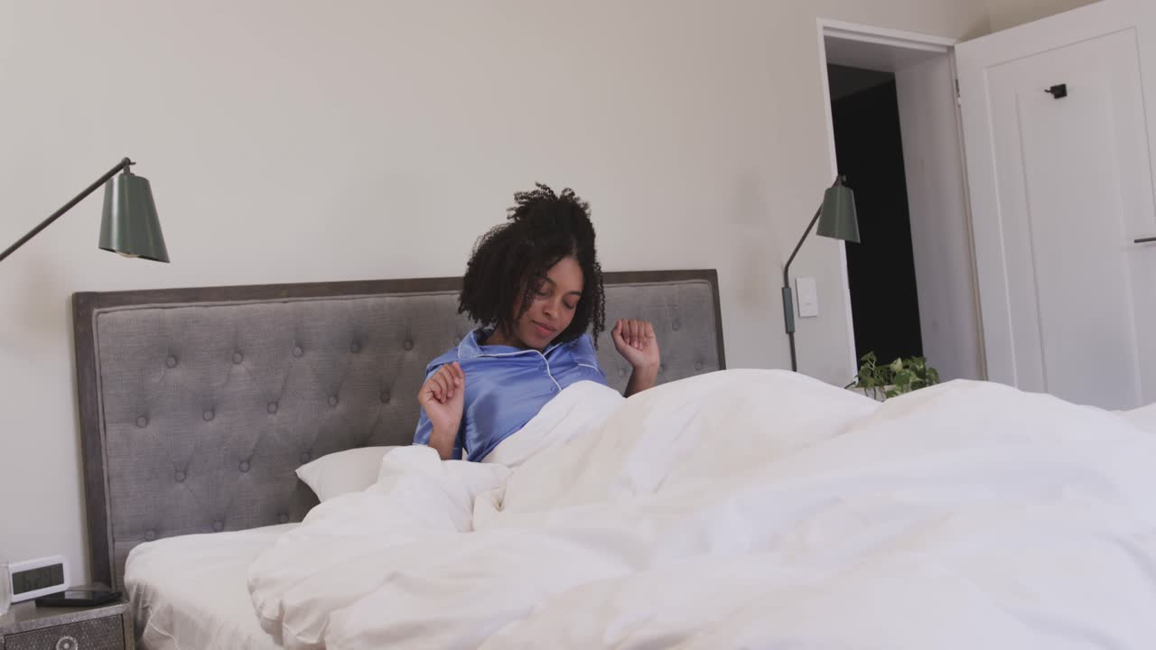 Donna afroamericana che si sveglia con la sveglia, si siede e si stira vestita in camera da letto, iniziando la giornata.