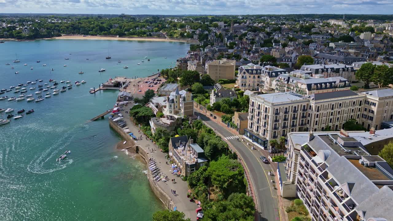 Drone flies along Dinard coastline, showing marina, turning boat, bay full of vessels, road, beach, and residential buildings