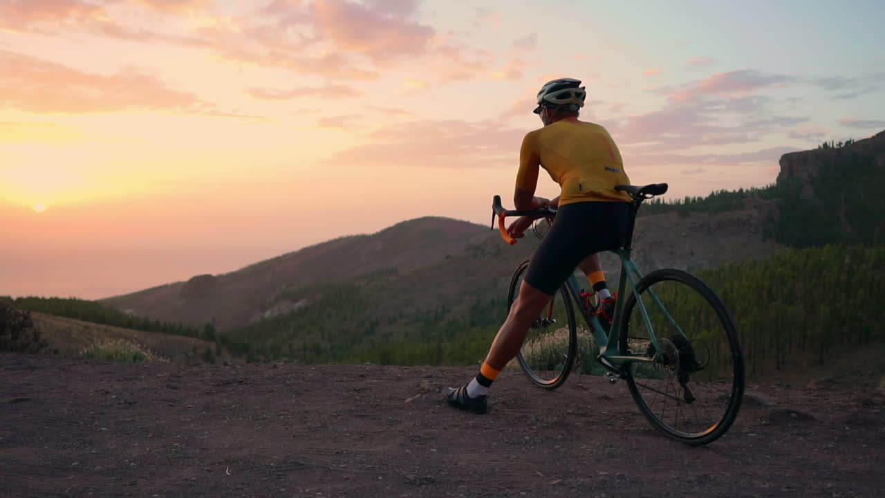 With mountains and sunset in view, sports enthusiast in yellow t-shirt and gear takes a break on a bike at mountain's peak after training