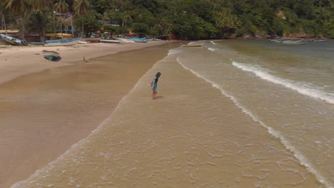 A-girl-in-a-blue-dress-playing-in-the-waves-crashing-at-shoreline-with-fishing-boats-and-a-village-in-the-background