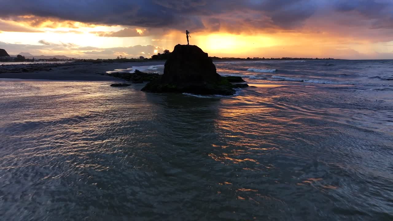 Sunset waves crash against a rocky outcrop, spray rising around a lone statue silhouetted in gold—still and solemn amid the ocean’s restless glow.