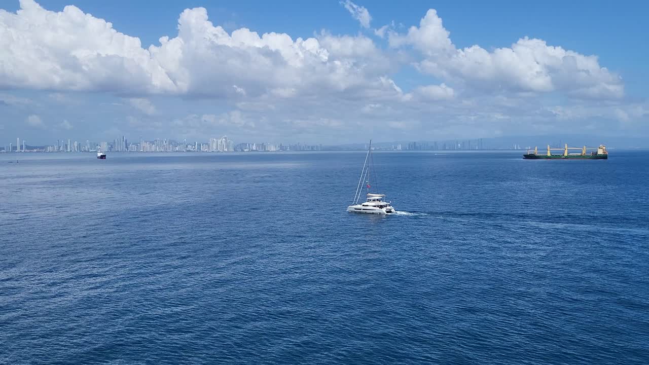 A white catamaran and a cargo ship sail across calm blue ocean waters with Panama City skyline in the background.