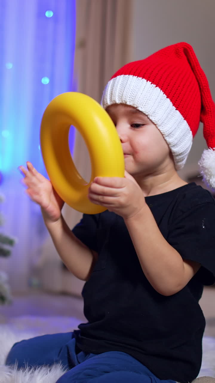 Adorable two year old boy in Santa cap rubs his eyes. Kid plays with toy near the Christmas tree. Vertical video
