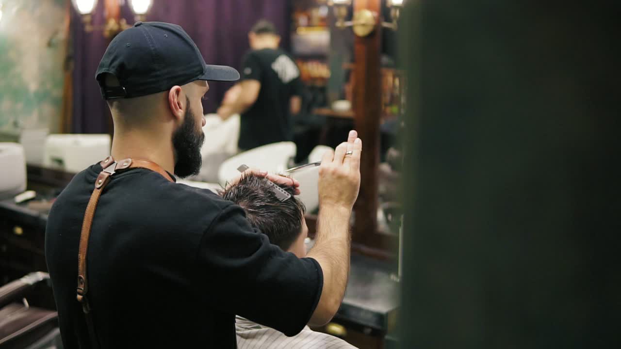 Side and back view of the stylish barber with beard performing a haircut with scissors holding a hairbrush in his hands. Slow
