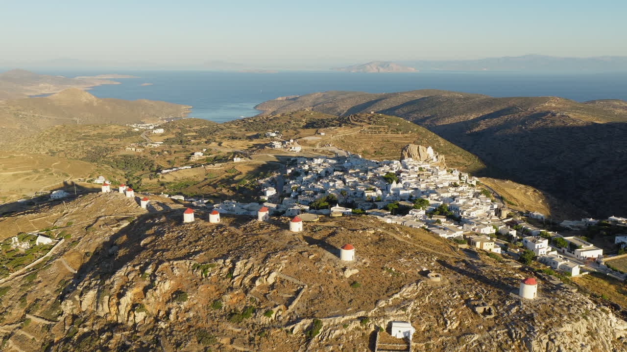 Slow panoramic drone orbit of Chora, Amorgos at sunrise, showcasing the village’s iconic white windmills glowing in the soft morning light