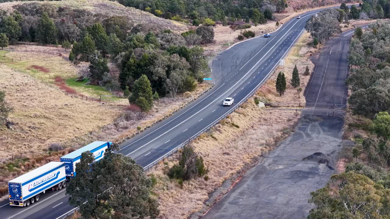Aerial footage captures a blue coach bus overtaking a white car on a winding rural road near Coonabarabran, Australia, under soft natural daylight