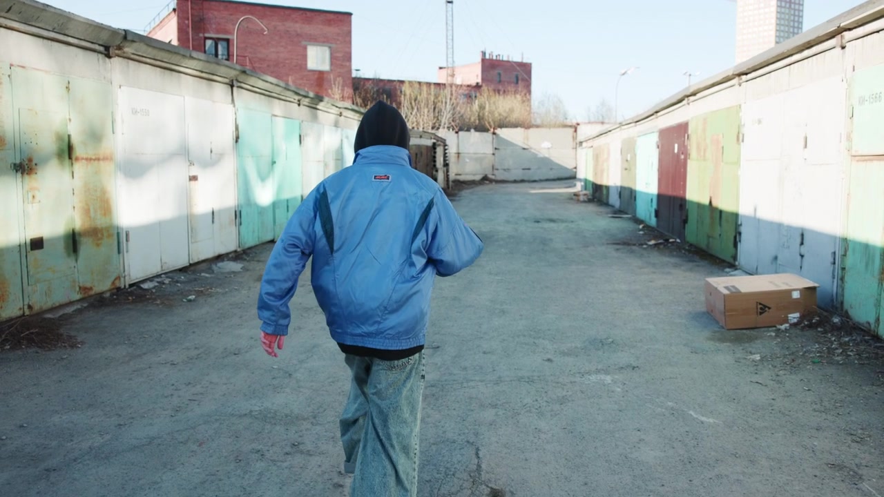 Una persona di spalle cammina lungo un lungo vicolo di deposito delimitato da porte di garage metalliche macchiate di ruggine e cemento grezzo, con luce diurna fredda, prospettiva centrata, superfici usurate e un'atmosfera urbana tranquilla.