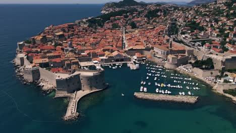 High-aerial-drone-footage-showing-entire-walled-city-of-Dubrovnik,-Croatia-circling-around-to-show-the-port-as-well-as-the-Dalmatian-coastline-landscape-in-the-distance