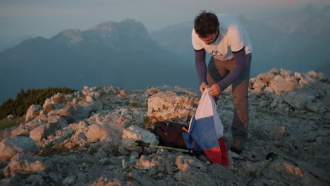 en la cima de la montaña peca un joven excursionista sacando una bandera eslovena de su mochila en la mañana del conde