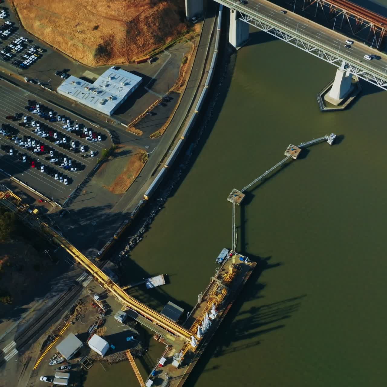 Train moving at the waterfront of California bay under Martinez bridge. Industrial area with docks and big parking lots. Top view