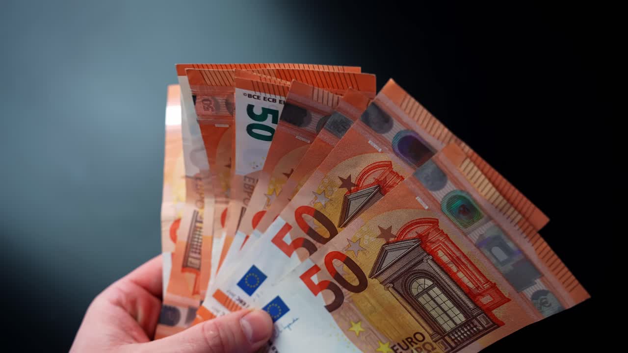 Metin ekleme alanı bulunan karanlık bir arka plan üzerinde bir el 50 avroluk banknotları yelpaze gibi savuruyor. Nakit ödeme, maaş, bütçeleme, birikim, kar ve kişisel finans konsepti.