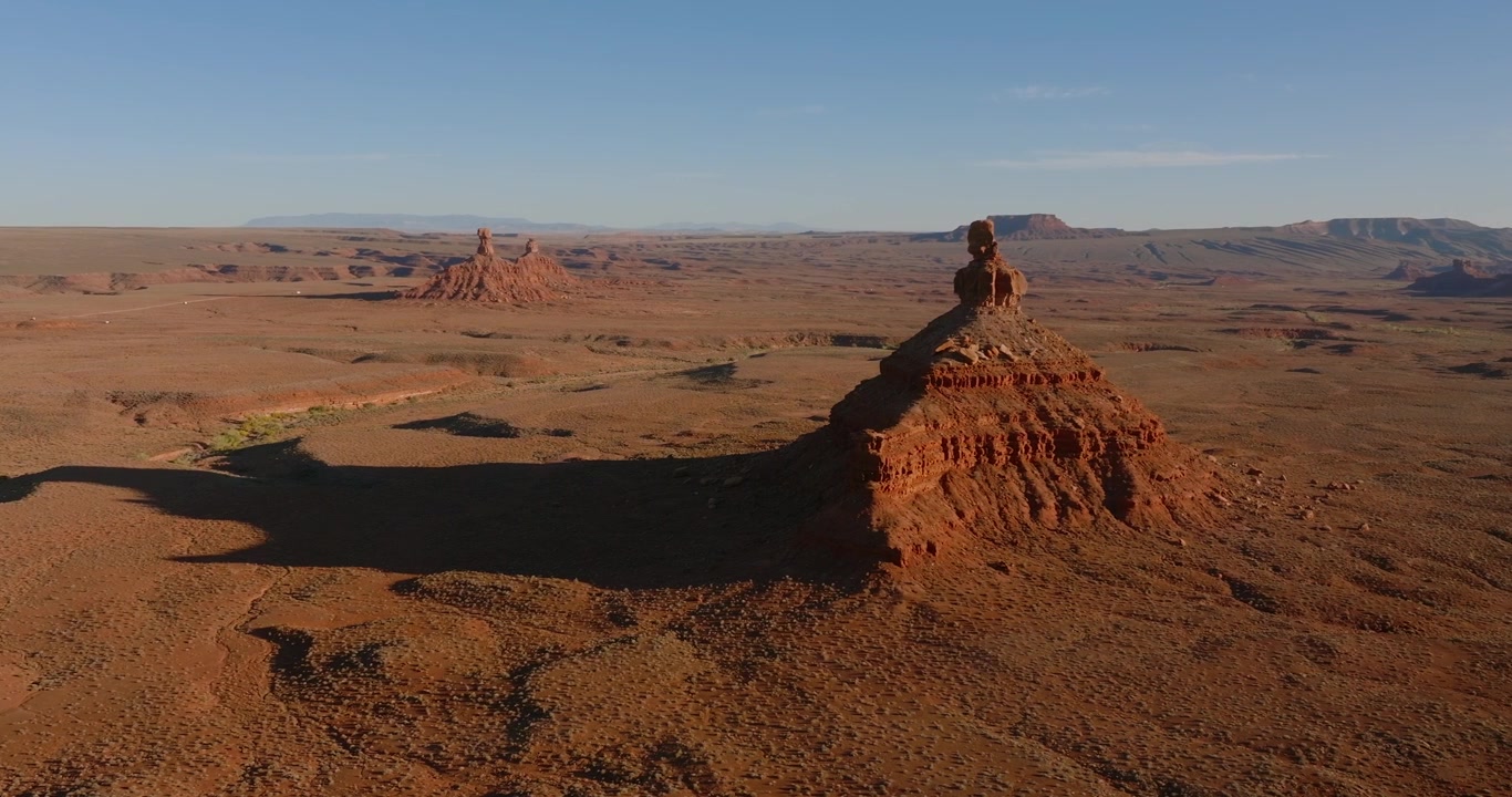 Il drone si muove lateralmente oltre Rooster Butte nella Valle degli Dei, Utah, rivelando un vasto paesaggio desertico rosso con formazioni e montagne distanti sotto cieli sereni al tramonto.