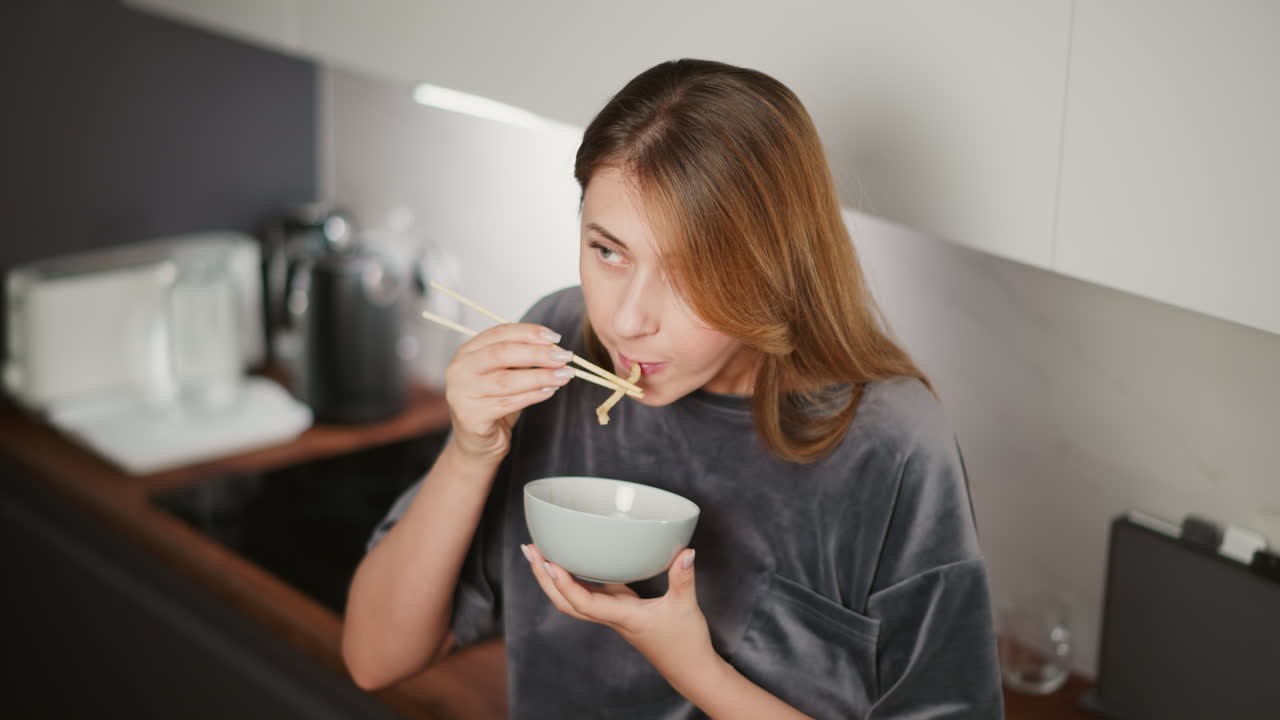 Young house girl with long hair eating lunch with chopsticks while resting casually against kitchen cabinet in modern kitchen, blurred view of glass cup and jug on wooden countertop