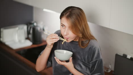 young house girl with long hair eating lunch with chopsticks while resting casually against kitchen cabinet in modern kitchen, blurred view of glass cup and jug on wooden countertop