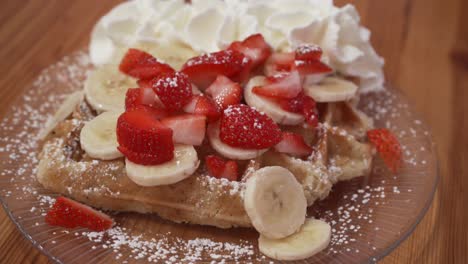 slow-upwards-moving-pan-of-belgium-waffles-with-whipped-cream-strawberries-and-banana-slices,-delicious-sweet-dessert-on-wooden-table