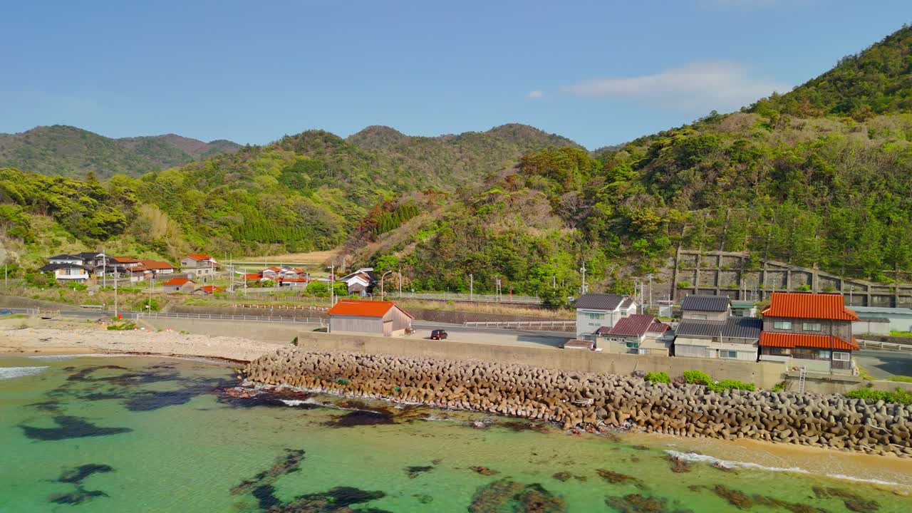 An aerial shot pushes in towards a quiet Japanese village on the coast, nestled between the clear turquoise sea and vibrant green mountains