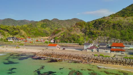 an aerial shot pushes in towards a quiet japanese village on the coast, nestled between the clear turquoise sea and vibrant green mountains