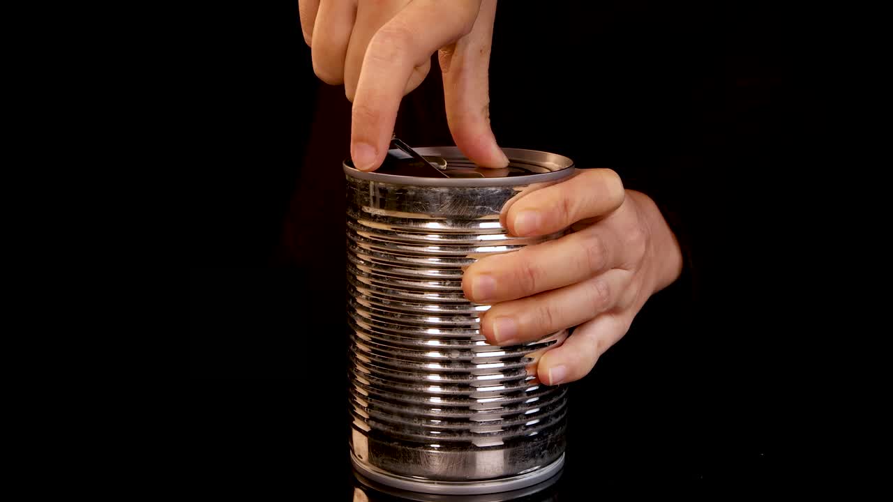 A human hand methodically opens a metal can of food, pulling back the lid under studio lighting with a dark, uncluttered background and steady camera