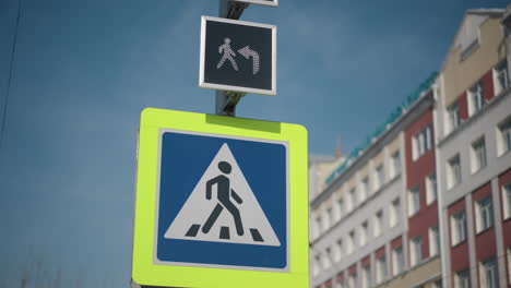 close view pedestrian crossing sign on pole under bright winter sun, luminous yellow frame, blue triangle symbol, city buildings behind, clear sky, urban safety marker for walkers and drivers