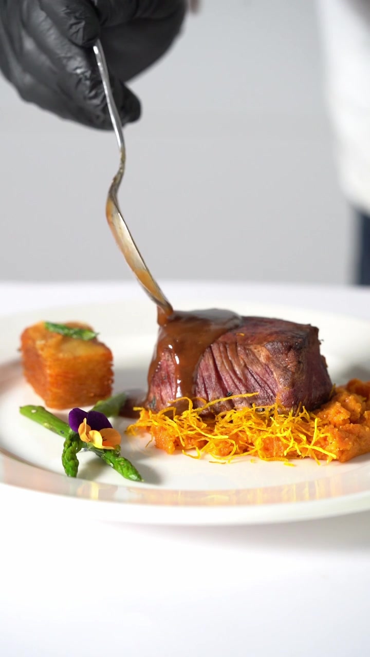 A chief puts sauce on a Juicy grilled beef steak served on creamy mashed vegetables in a minimalist plate, close up shot