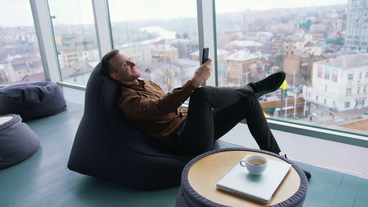 Young businessman relaxing near the window. Handsome man sitting in a cosy chair and having a video conversation with the city window view. Profile view.