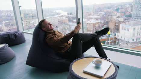 young businessman relaxing near the window. handsome man sitting in a cosy chair and having a video conversation with the city window view. profile view.