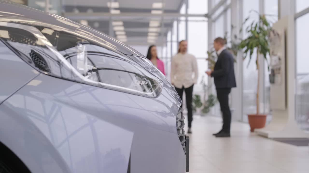 젊은 자동차 판매자가 대리점 살롱에서 젊은 부부에게 새로운 자동차를 보여주고 있습니다.