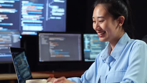 close up side view of asian female programmer drinking coffee while writing code by a laptop using multiple monitors showing database on terminal window desktops in the office