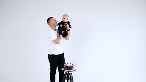 happy caucasian dad holding a little baby in black suit. father and son stand near the stool with a chocolate cake. white backdrop.