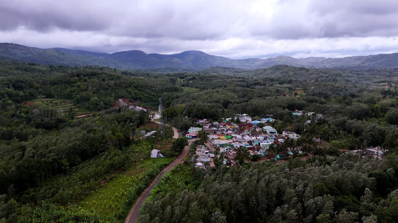 Drone view of a secluded settlement amidst rolling, tree-covered mountains. The scene is moody with an overcast sky, capturing the beauty of a remote, tropical hillside