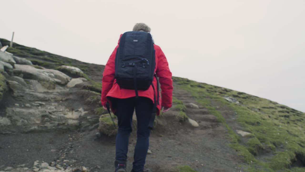 hombre caucásico con chaqueta roja con mochila haciendo autostop en una colina de montaña en las islas feroe, la cámara sigue desde atrás en un día nublado en cámara lenta