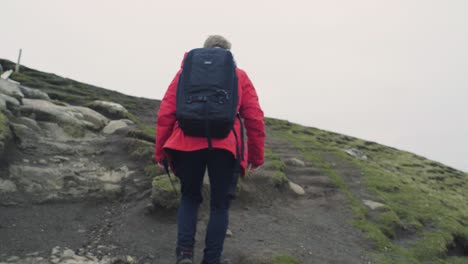 Caucasian-male-in-red-jacket-with-backpack-on-hitchhiking-up-a-mountain-hill-on-the-Faroe-Islands-camera-follows-from-behind-on-a-cloudy-day-in-slow-motion