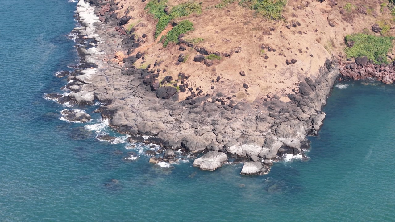 Aerial drone view of a rugged rocky headland meeting the clear turquoise water of the Arabian Sea at Mithmumbari Beach, Maharashtra, India.