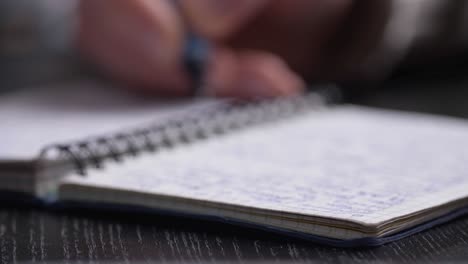 close up of man's hands writing in spiral notepad placed on wooden black desktop