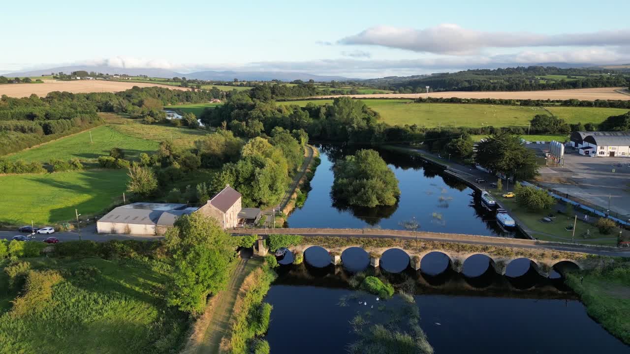 Vue aérienne par drone d'un pont en arc de pierre traversant une rivière calme en Irlande rurale, montrant des terres agricoles verdoyantes, des arbres et une campagne paisible sous une douce lumière du soir.