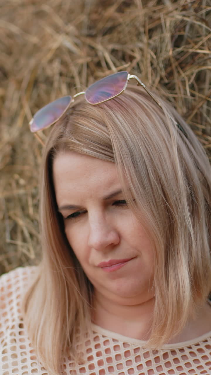 mujer apoyada en una bala de heno, con gafas de sol en la cabeza y una camiseta de tela neta, un entorno rural pacífico con hierba seca, cielo azul y expresión relajada