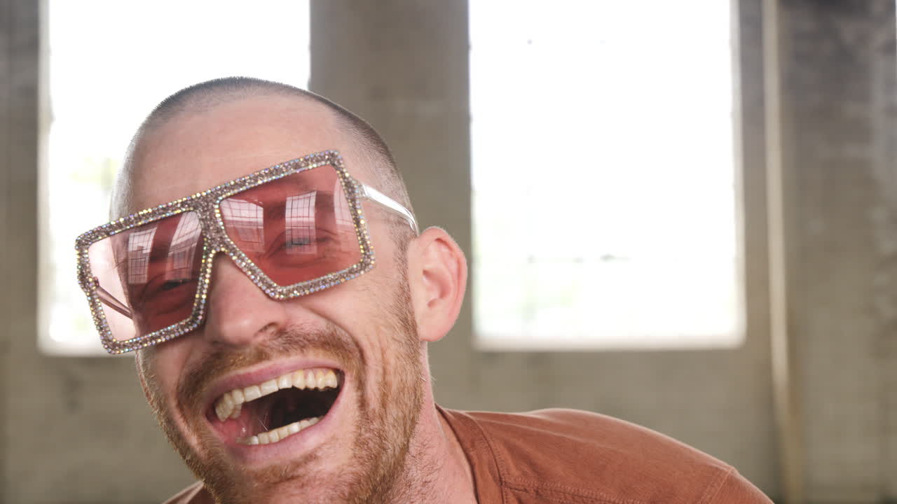 Slow motion footage of a man in an industrial warehouse setting wearing rose tinted glasses laughing into camera.
