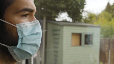 hombre mirando por una ventana ansiosamente poniéndose una máscara facial en cuarentena de cerca