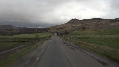 Toma-En-Movimiento-De-Un-Vehículo-Que-Conduce-Por-La-Pequeña-Carretera-Con-Una-Luz-Dramática-Y-Nubes-Y-Montañas-En-Edimburgo,-Escocia