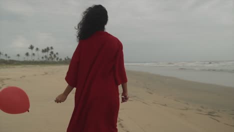 Una-Hermosa-Mujer-Con-Un-Vestido-Rojo-Sosteniendo-Un-Globo-Rojo-En-La-Playa-Mirando-Hacia-El-Océano,-Una-Mujer-India-Con-Globos