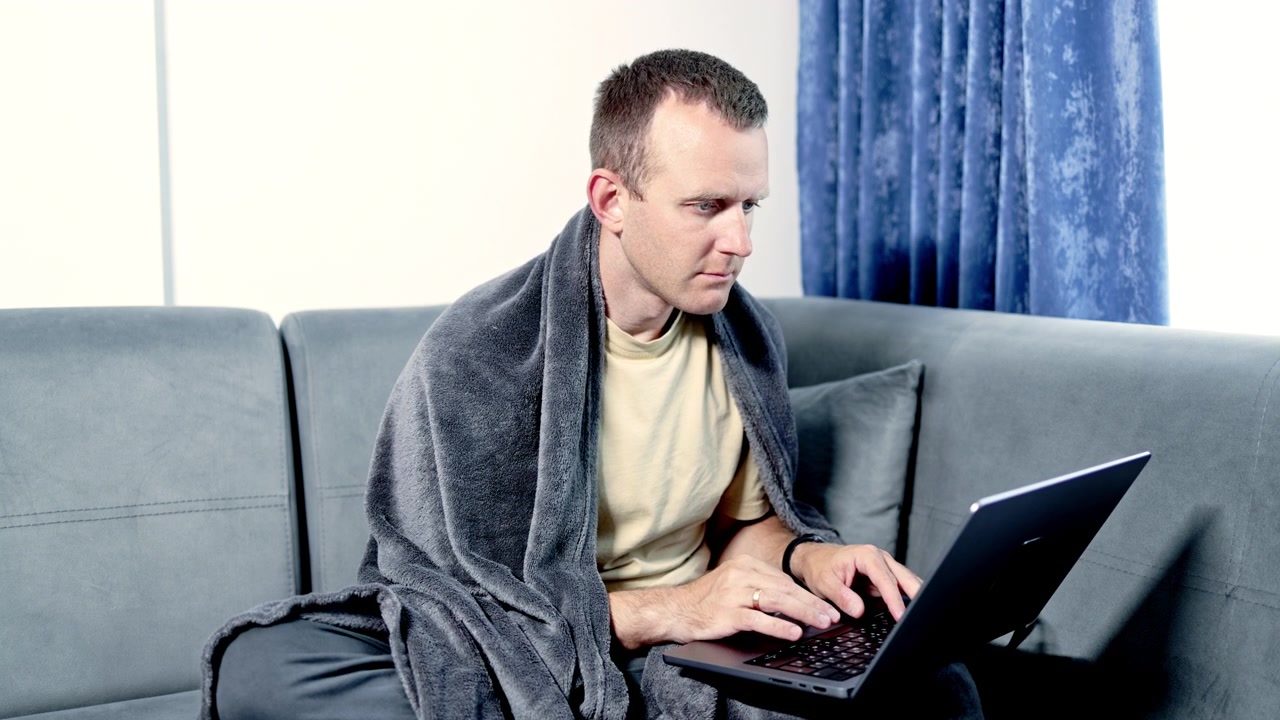 Uomo seduto sul divano che usa un laptop, avvolto in una coperta, di giorno in uno spazio interno accogliente