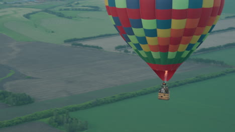 colorful checkered hot air balloon carrying passengers drifting above patchwork field at sunset with visible flame jet, serene aerial motion over green fields and distant horizon under sky clouds