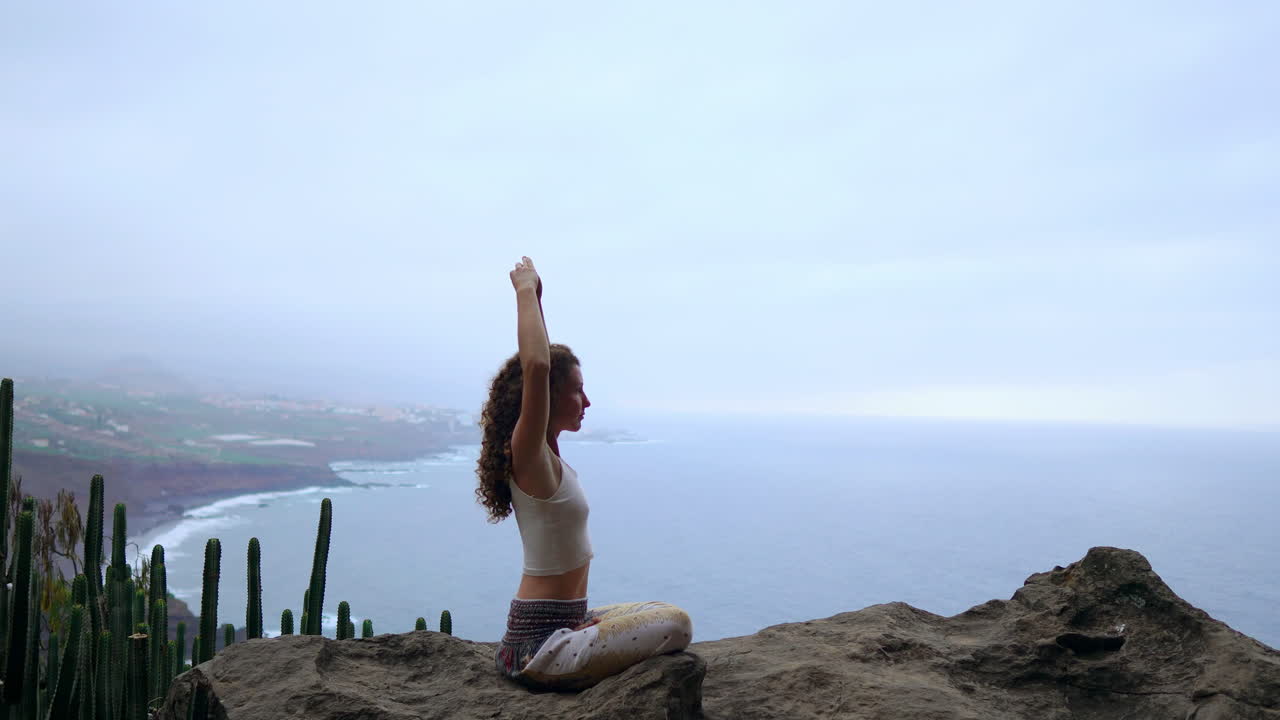 On a mountain peak, a woman meditates and shapes the Maha Sakal hand gesture, with the ocean and verdant mountains enhancing her ambiance