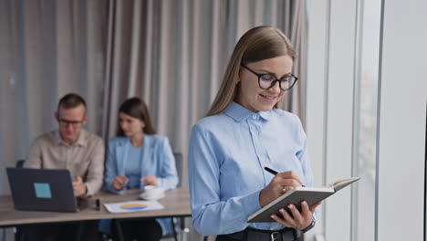 pretty smiling lady wearing glasses writing something in her notebook. a pair of colleagues talk sitting in front of laptop at backdrop.