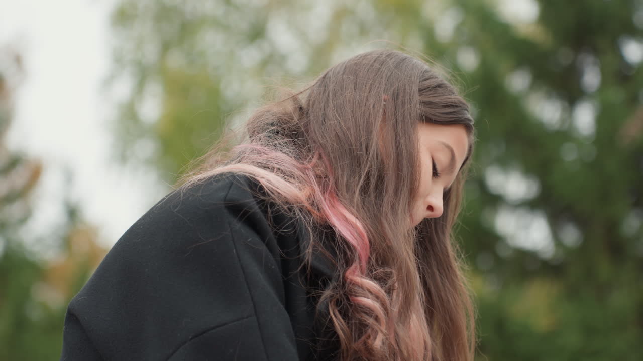 Garota perdida em seus pensamentos em um banco de parque. Jovem garota ponderando sob o céu nublado em um ambiente de parque tranquilo. Garota contemplativa com cabelo rosa sentada silenciosamente em um banco sob nuvens cinzentas