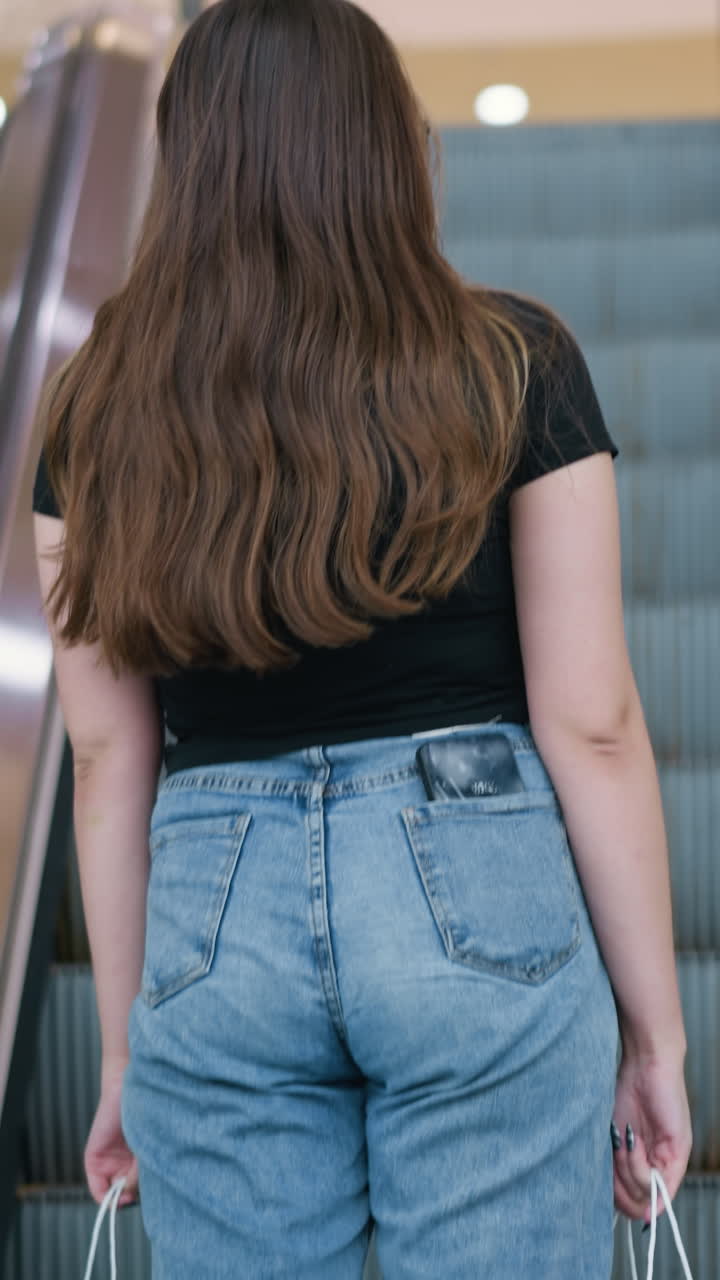 검은 티셔츠와 파란 청바지를 입은 긴 갈색 머리카락을 가진 젊은 여성이 현대 쇼핑몰의 에스컬레이터에 올라 밝은 조명 아래 양손으로 쇼핑 가방을 들고 있습니다.