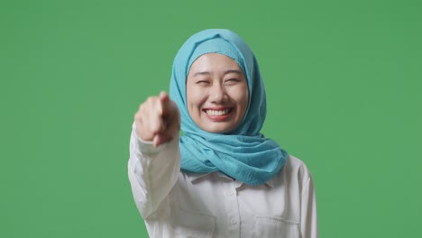 아시아 무슬림 여성이 미소 짓고 가을 만지며 녹색 스크린 배경 스튜디오에 서있는 동안 당신을 가리키고 있습니다.