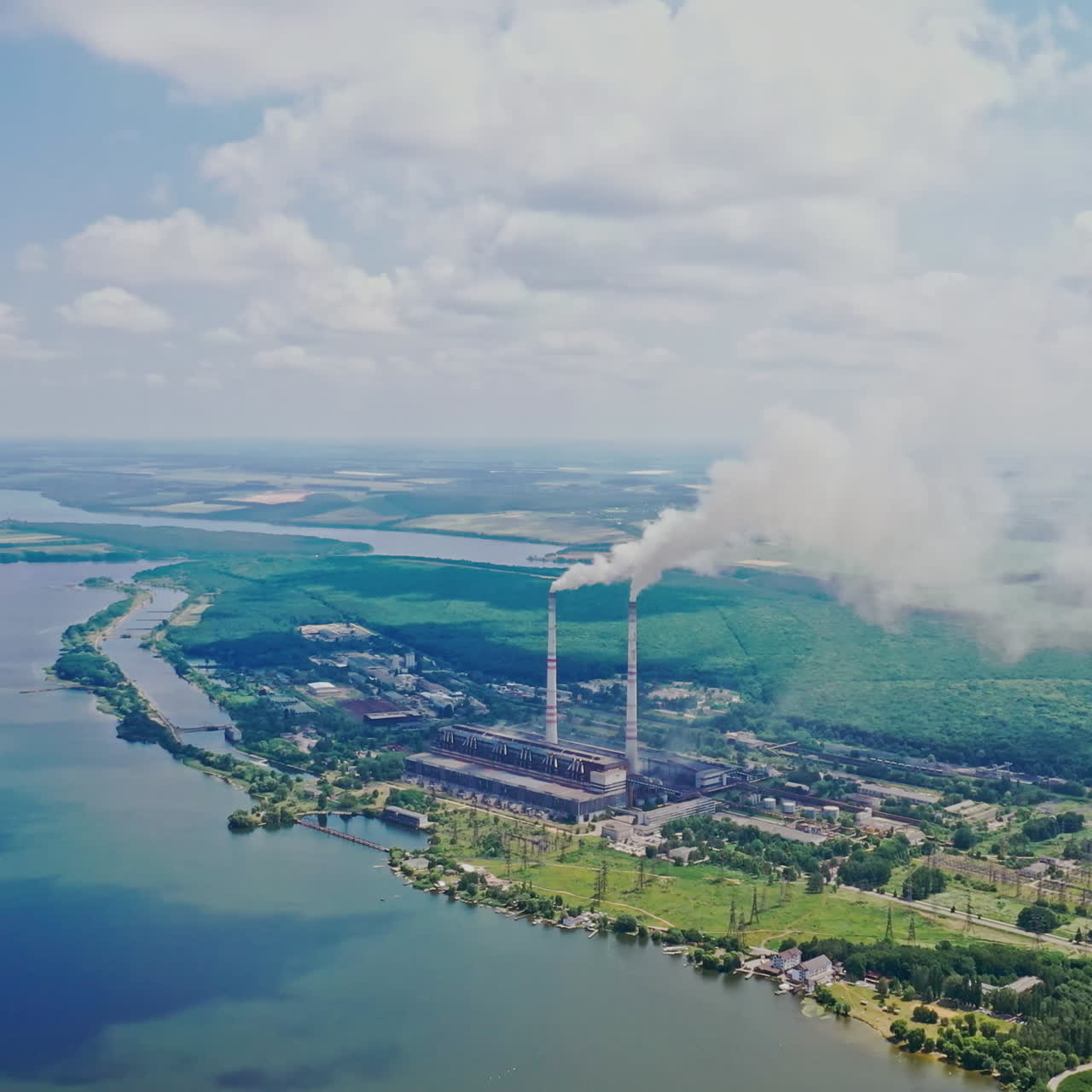 Huge manufactory plant among natural background. Smoke comes from pipes into the atmosphere near the river and green forest outdoors. Aerial view.